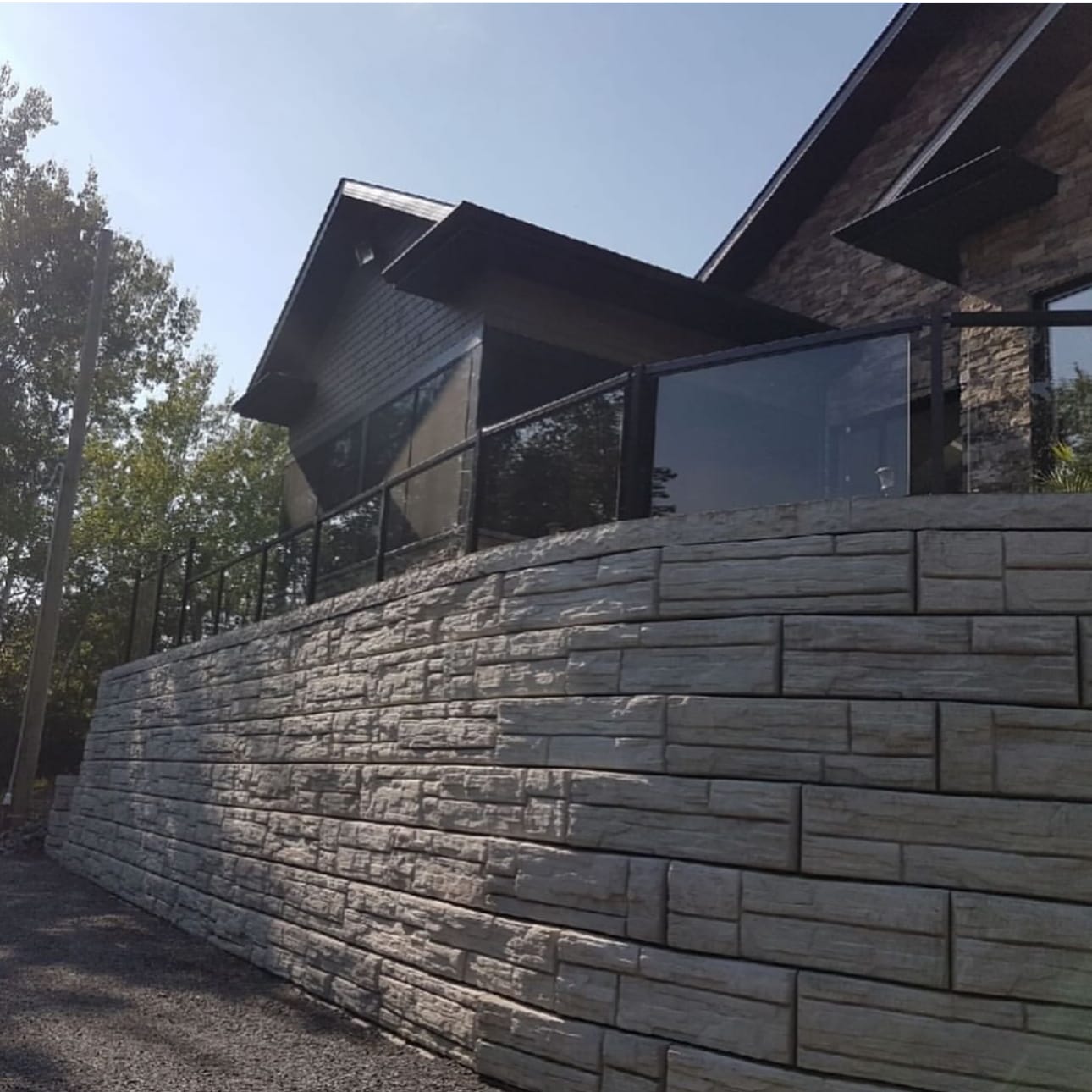 Tall stone-look block retaining wall below a house, with a black-framed glass railing along the top