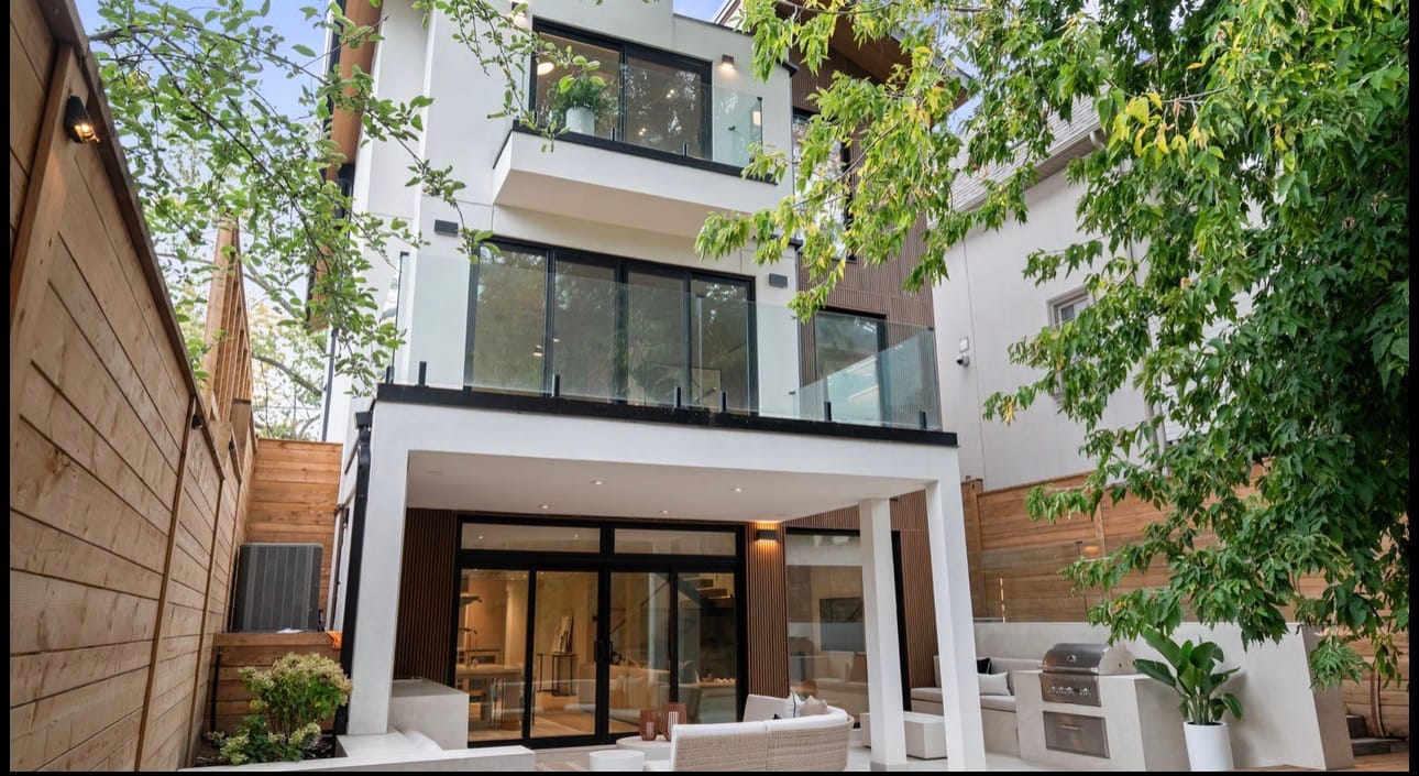 Covered rear patio with an outdoor kitchen and seating under glass-railed balconies, beside a horizontal wood privacy fence