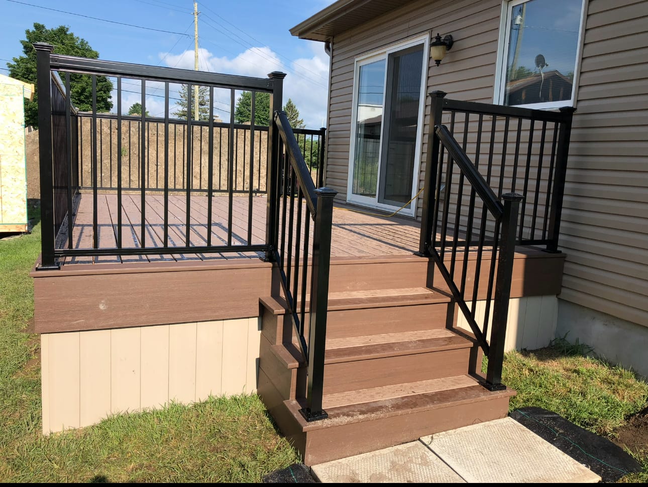 Small brown deck with black metal railings and steps down to a concrete pad, beside a house with tan siding