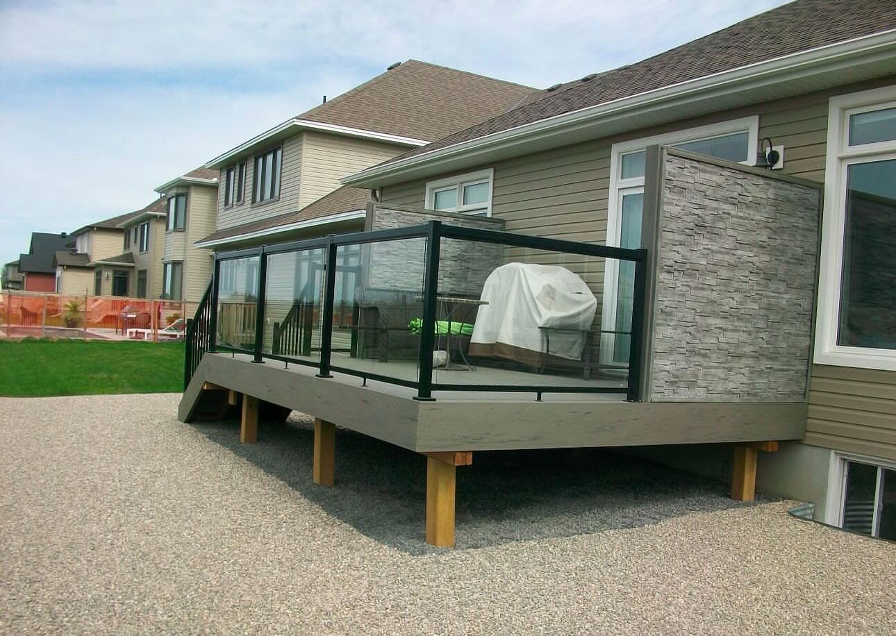 Raised grey deck on wood posts with a black-framed glass railing and a stone-look privacy wall, over a gravel yard