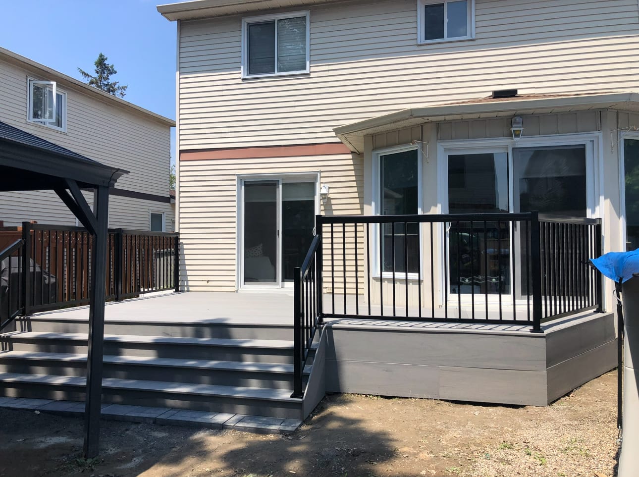 Low grey deck with full-width steps on one side and a black metal railing on the other, behind a house with beige siding
