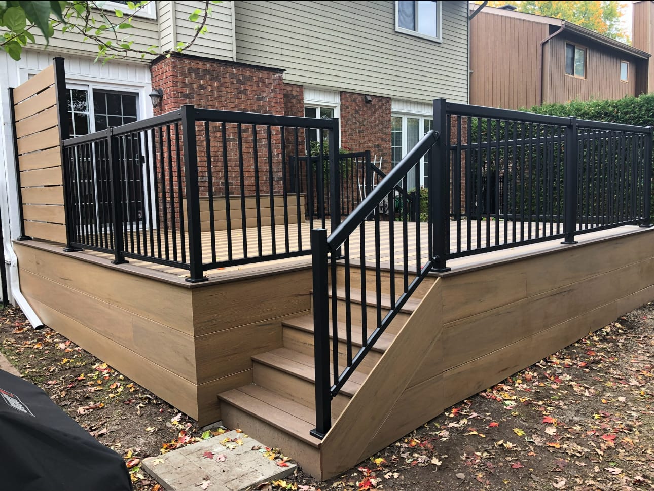 Tan deck with board-clad skirting, black metal railings, stairs and a horizontal privacy screen at one end