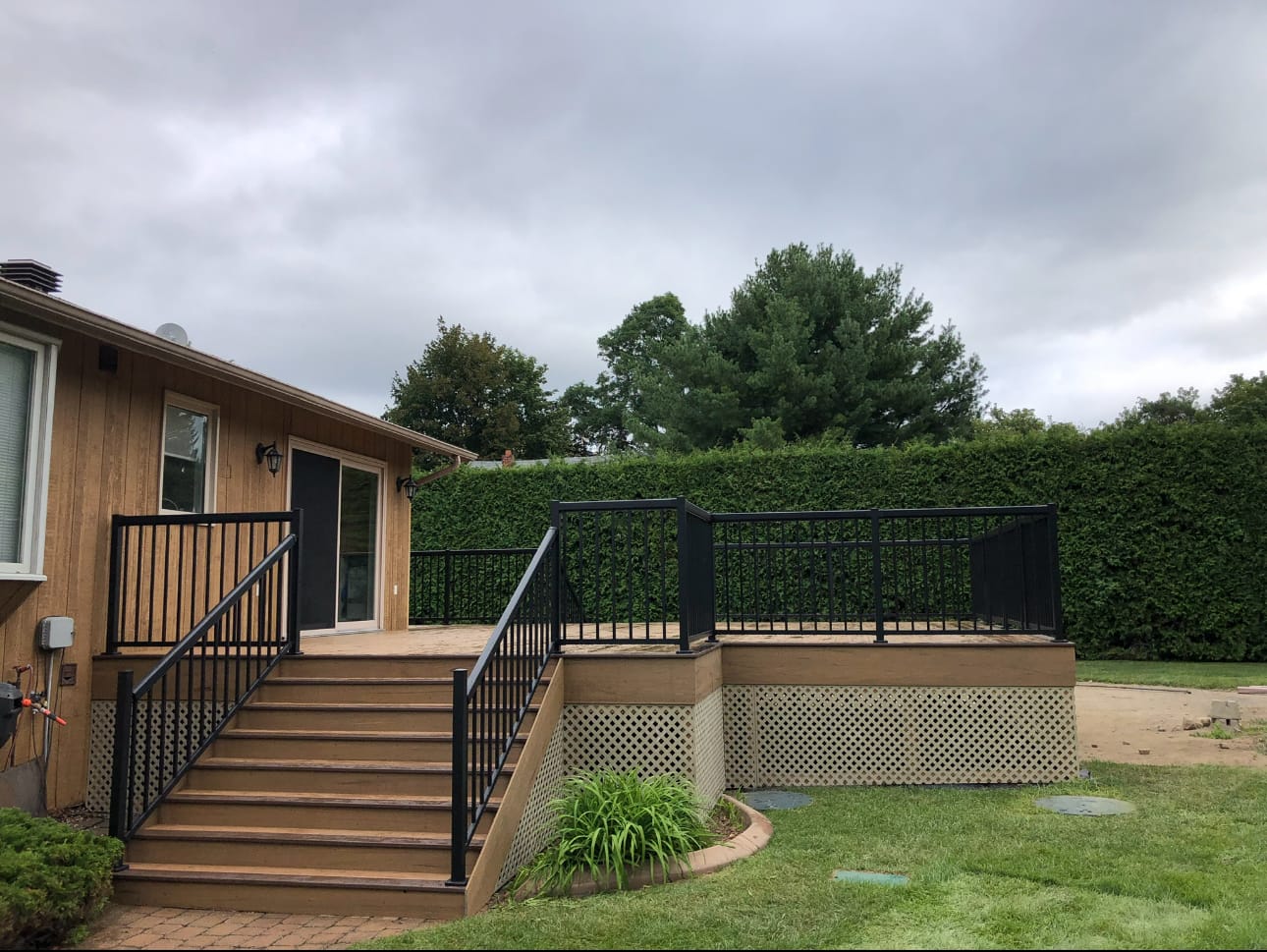 Brown deck with wide stairs, black metal railings and lattice skirting, in front of a tall hedge
