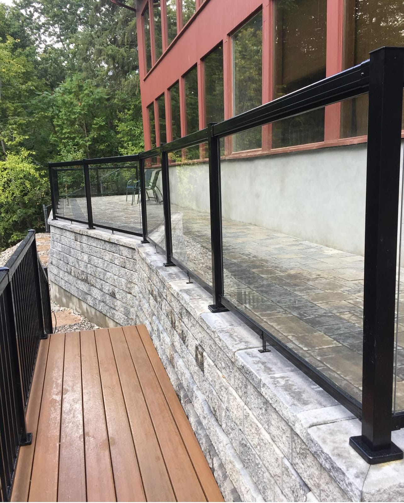 Black-framed glass railing along the top of a block retaining wall above a stone patio, with a brown deck walkway below