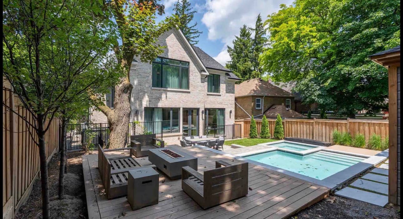 Low deck with a fire table and lounge seating beside a pool and spa, inside a wood privacy fence