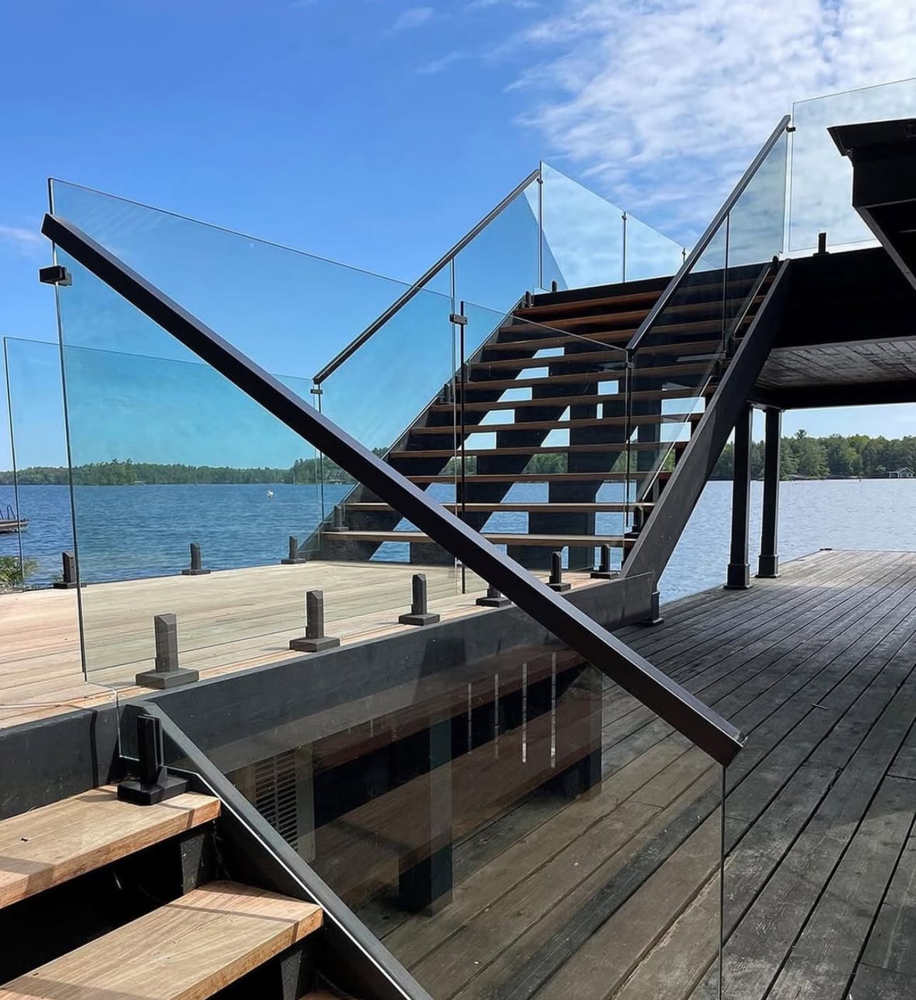 Open-riser wood stairs with a glass railing rising from a waterfront deck to an upper level