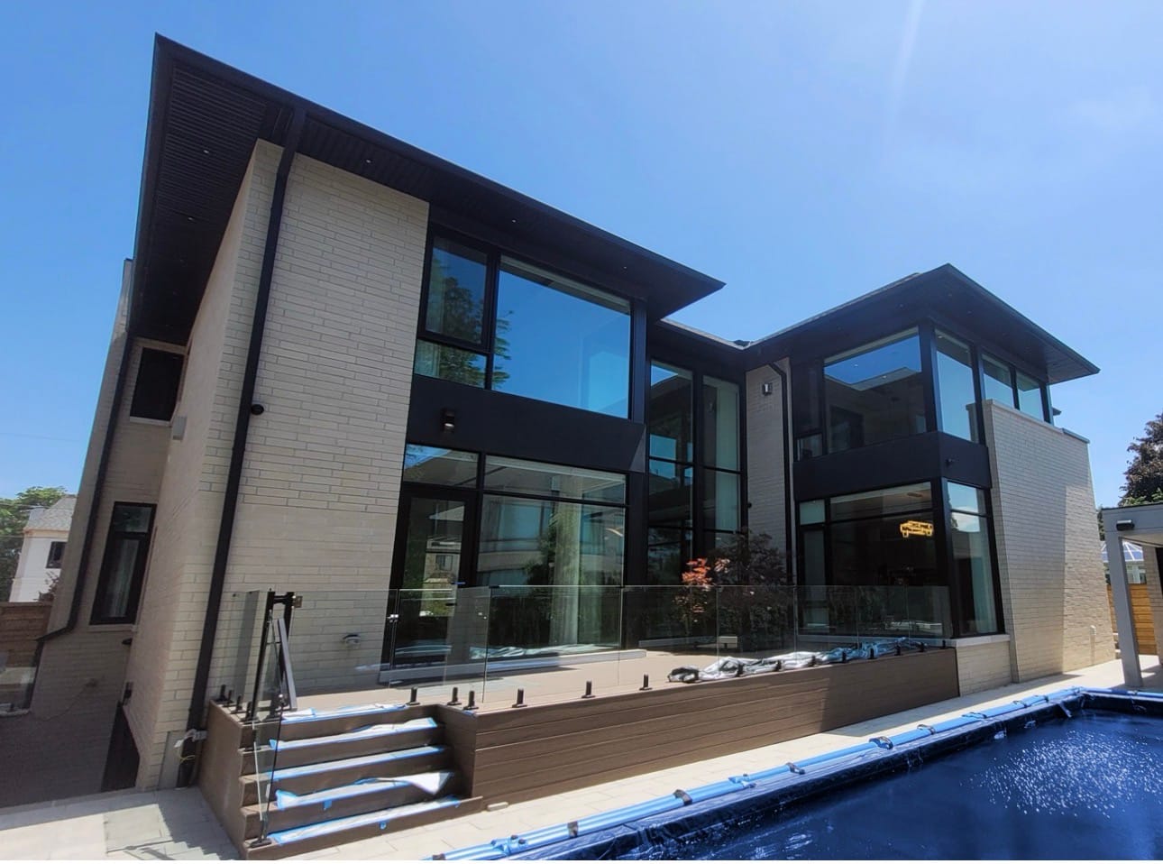 Raised brown deck with a glass railing and steps down to a pool, behind a modern house