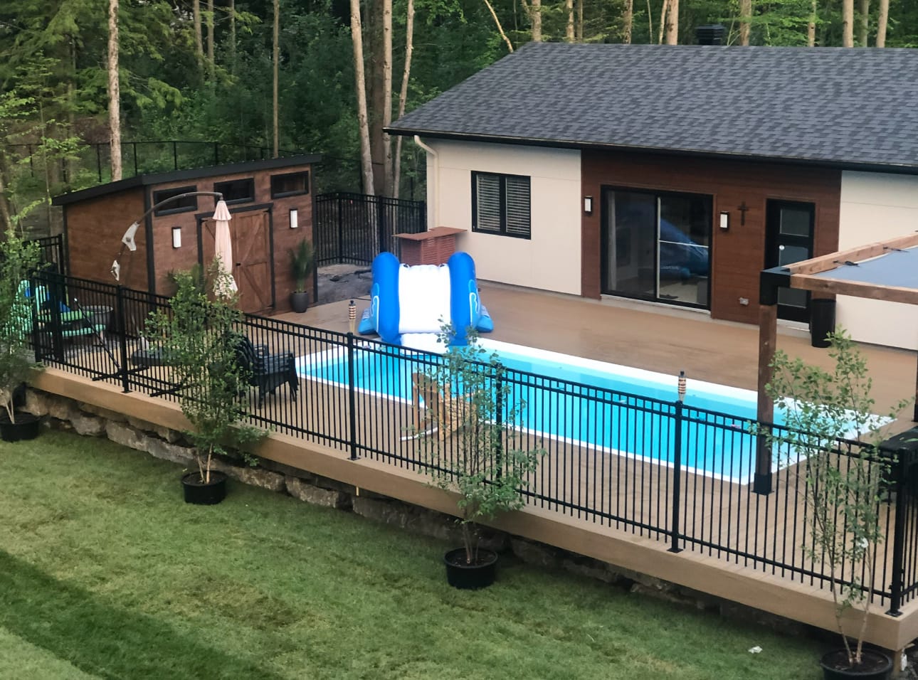 Black metal picket fence along a brown deck that surrounds an in-ground pool, with a shed, a pergola and a pool house behind