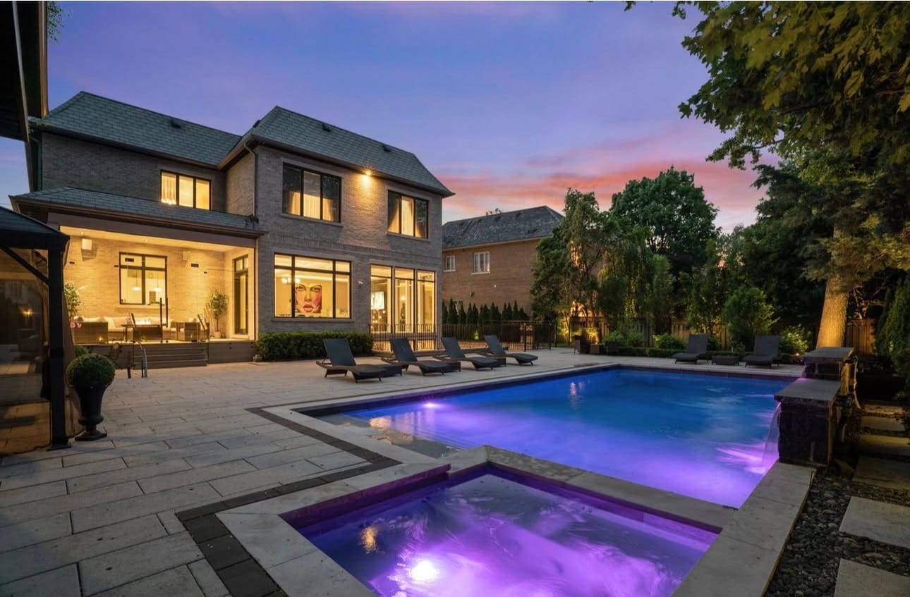 Pool and spa lit at dusk, surrounded by large-format stone paving behind a brick house