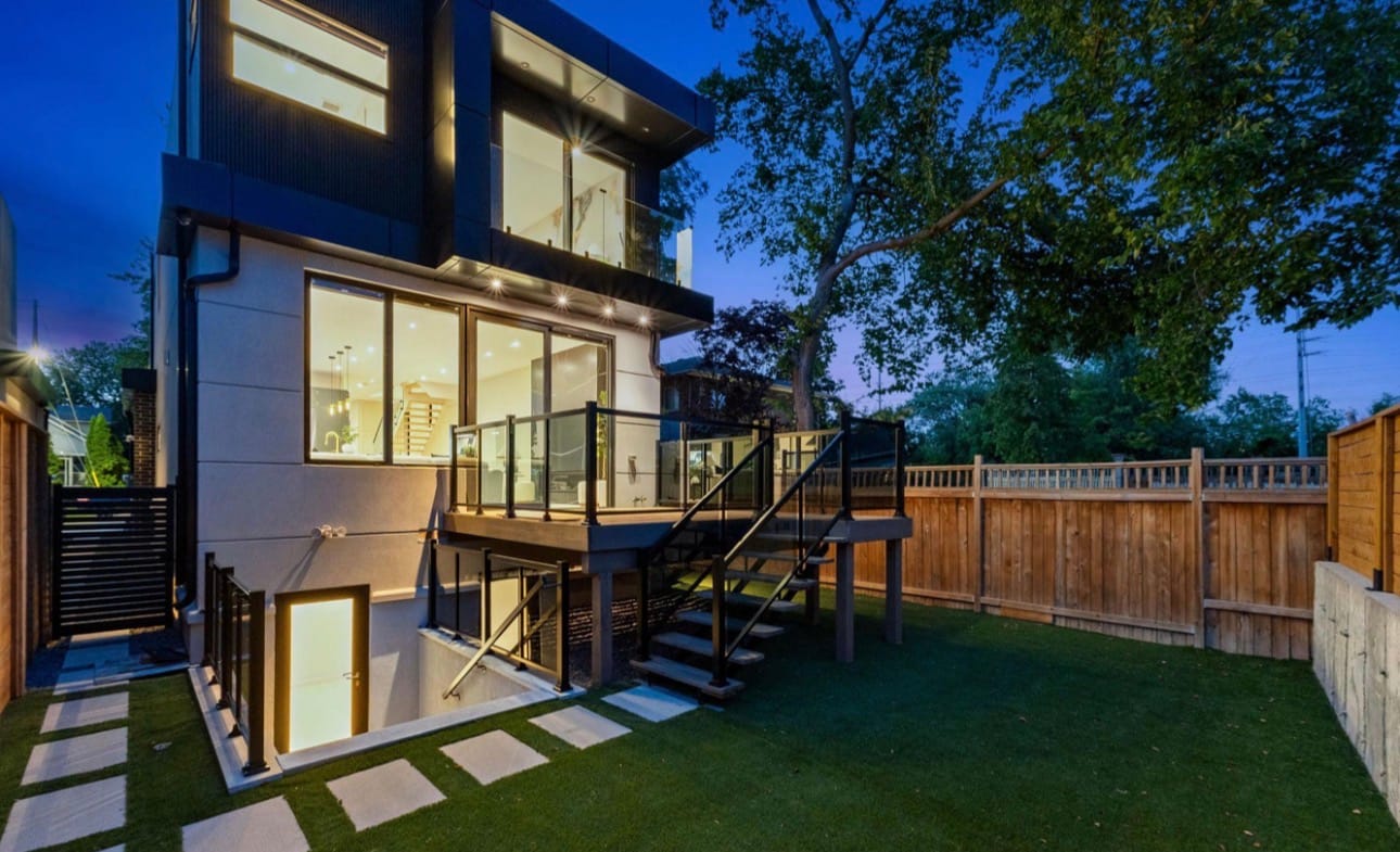 Raised rear deck with a glass railing and open stairs down to a small yard of turf and stepping stones, at dusk
