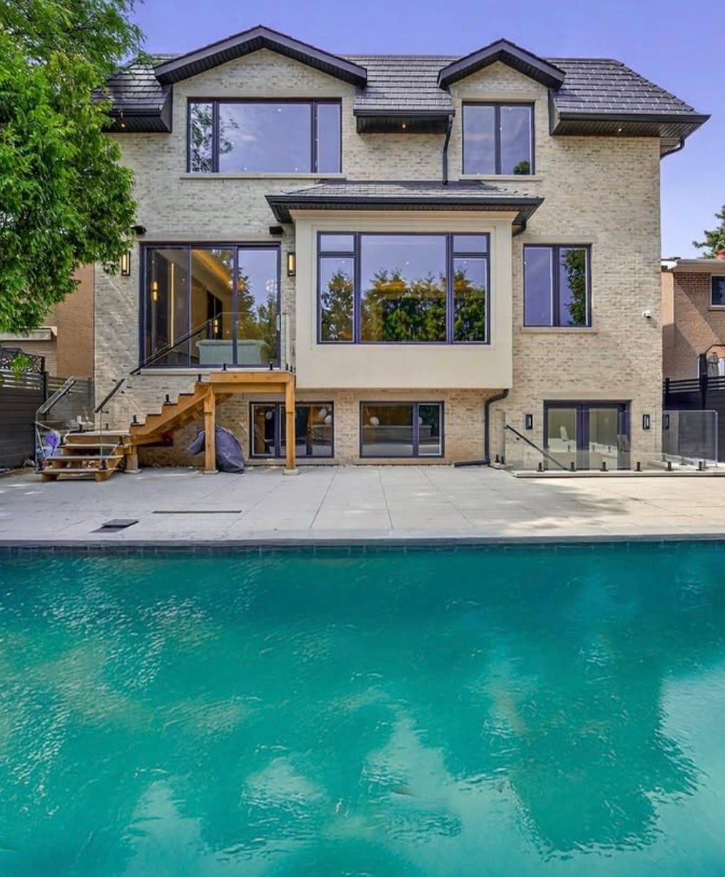 Wood stairs and a raised deck at the back of a house, seen across a pool, with glass railing at the lower walkout